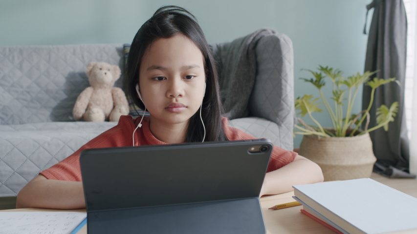 Asian girl is studying online via internet on tablet with headphone while sitting at home morning and sunlight. Asia children using digital computer and write homework. Concept online learning at home Royalty-Free Stock Footage #1053684749