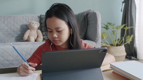 Asian girl is studying online via internet on tablet with headphone while sitting at home morning and sunlight. Asia children using digital computer and write homework. Concept online learning at home