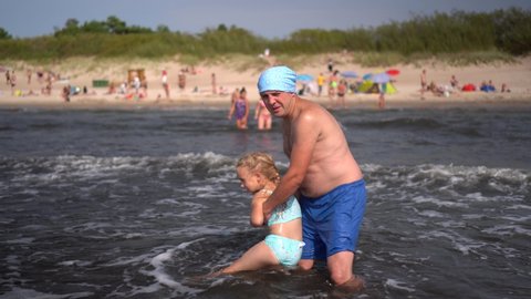 Playful daddy father with headscarf turn wash his cute daughter in sea waves water. Loving man with little girl having fun. Blurred people on beach in background. Camera motion shot with gimbal.