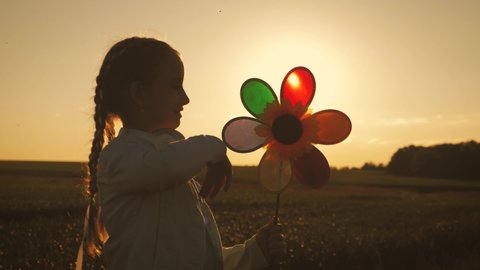 Cheerful girl are running in the meadow with wind turbines as a sign of freedom. Happy child holding wind toy running in green field, rejoicing life. Silhouette of kid girl holding wind toy.