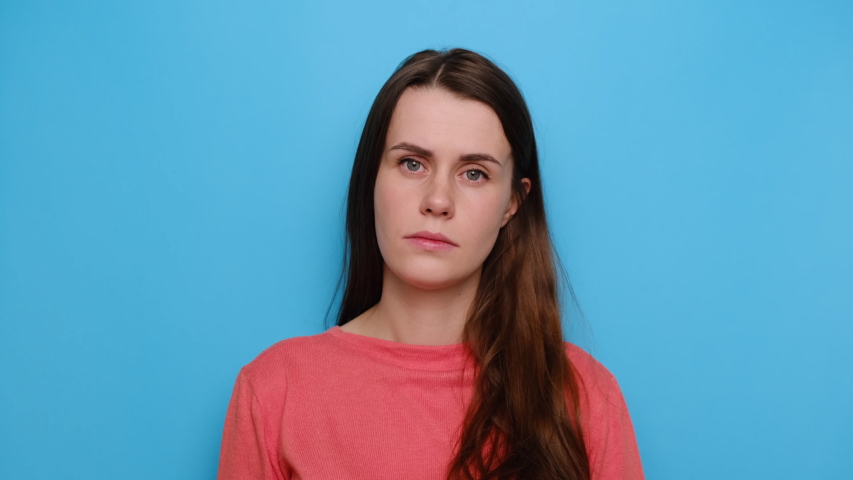 Exhausted young woman feeling bored, lack of motivation, apathy, wears sweater, isolated on blue studio background. Sad girl with frowning expression, looks unhappy straight at camera. Human emotions | Shutterstock HD Video #1053691865