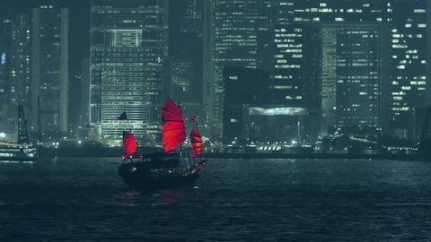 Hong Kong view at night from Victoria harbor with traditional red sail junk boat and modern skyscrapers and other urban buildings of financial district of city  