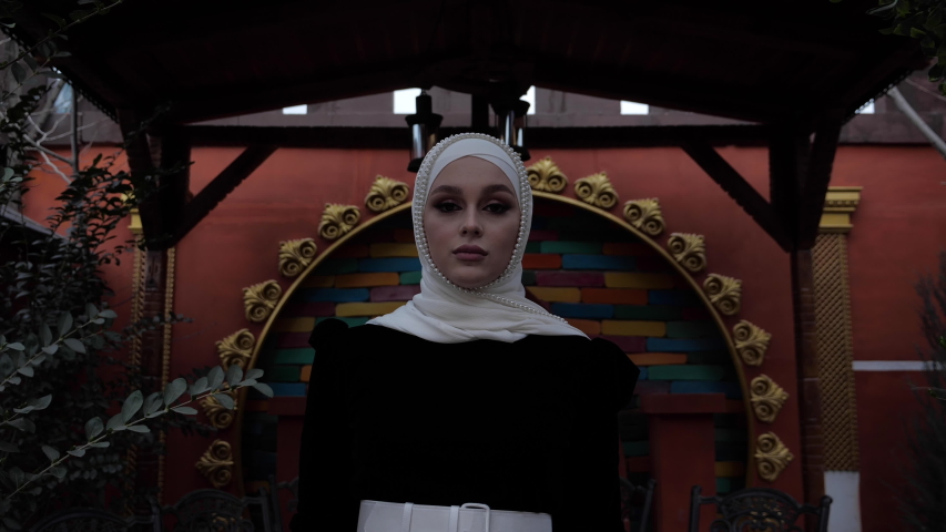 pretty woman in Arabian suit with white belt and hijab poses near green arch in national eco restaurant slow motion