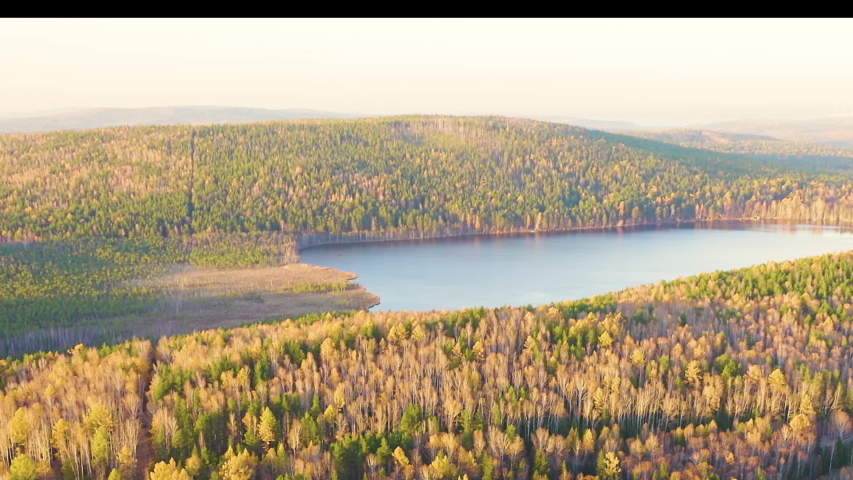 Lake Peschanoye (Sandy). A dense, mixed forest displaying peak autumn foliage surrounds the calm, blue expanse of Lake Peschanoye. The canopy presents a rich tapestry of vibrant golden-yellow birch and aspen leaves intermingled with the deep green of towering pines, creating a highly textured and multi-layered landscape. The lake is characterized by its sandy bottom and shores, which give the body of water its name, and a small rampart of granite blocks lines a section of the bank. To the west, an ancient swamp, over ten thousand years old, borders the lake. The camera executes a smooth, steady forward aerial movement, gliding over the treetops and gradually approaching the water