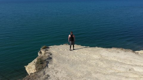 Aerial view of man stands on top of mountain near cliff. Traveler enjoys beautiful sea view.