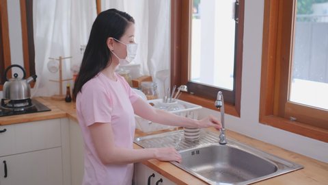 Beautiful woman wash hands in sink of home kitchen and also wear hygiene mask to prevent virus infection. Concept of new normal for good health lifestyle to protect from covid virus pandemic in city.