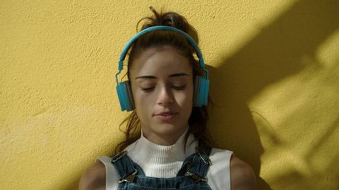 Attractive Young Woman stops and looks to the camera and smiles while listening to music wearing wireless headphones seated against a yellow wall on a sunny day medium shot zoom in