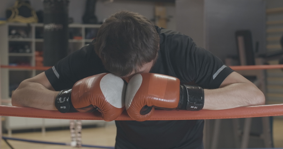 Portrait of tired handsome Caucasian boxer standing on boxing ring with hands on ropes. Exhausted bearded man raising head and sighing. Sport, exercising, training, lifestyle. Cinema 4k ProRes HQ.