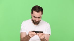 Gambling bearded young guy 20s in white t-shirt using play mobile cell phone gadget smartphone for video games isolated on green chroma key background studio. People sincere emotions lifestyle concept
