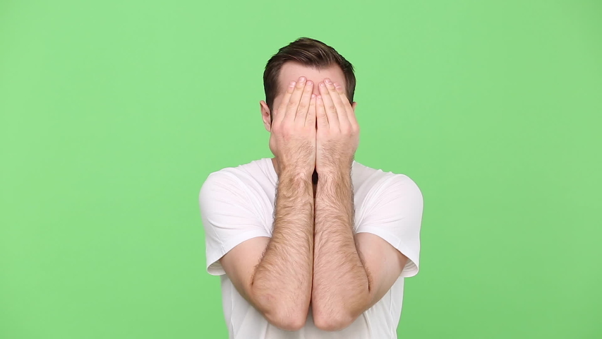 Bearded fun young guy 20s in white t-shirt isolated on green chroma key background studio. People sincere emotions lifestyle concept. Looking camera with scared fearful glance covered face with hands