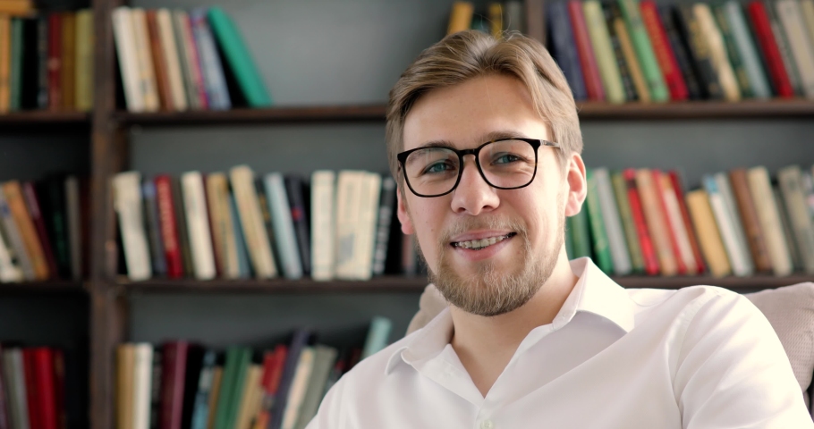 Portrait of young professor or student in glasses and white shirt at University library in campus looking at camera and smiling on bookshelves background. Working, studying, education concept. | Shutterstock HD Video #1053742529