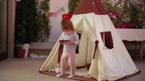 cute little toddler baby playing in toy wigwam, watching video on digital device