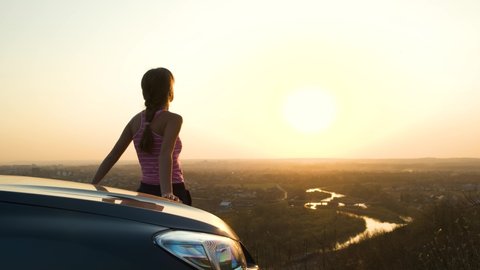 Young woman standing near her car enjoying warm sunset view. Girl traveler leaning on vehicle hood looking at evening horizon.