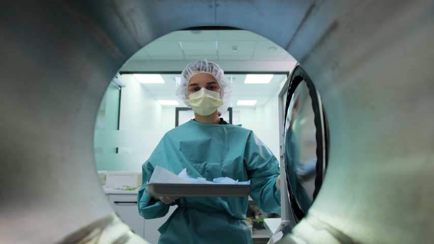 Lab nurse in green uniform, yellow mask and white cap puts pockets with dental tools inside autoclave sterilizer machine for disinfection. POV medical oven. Modern professional laboratory equipment. | Shutterstock HD Video #1053754979