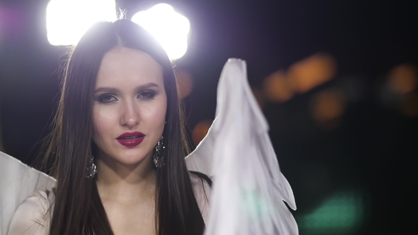 Beautiful girl angel costume with white wing closeup looking at camera sensual sight. Model woman angel suite fashion show sparkle slow motion. Female portrait emotion smile on face pretty dress.