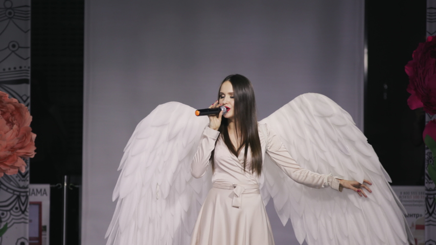Singer woman angel suite fashion show sparkle hold mic slow motion. Beautiful girl angel costume with white wing closeup sing song sensual sight 4K. Female portrait emotion smile on face pretty dress.