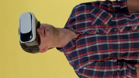 Dark-haired man using virtual reality glasses while standing against yellow background. He is in checkered shirt. Video with Vertical Screen Orientation 9:16
