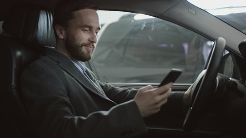 Surfing the internet happy handsome man sitting in the car on a driver's seat and using his phone. Businessman reding news while sitting in a car.