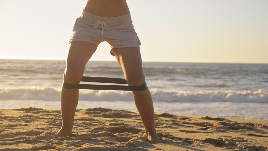Medium shot young successful athletic woman is doing exercise with elastic on her legs in sportswear on the ocean beach at sunset in orange glow. lifestyle fitness | Shutterstock HD Video #1053760181