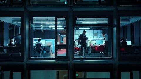Footage from Outside the Window: Businessmen and Businesswomen Working in the Office. Managers and Specialists Doing Financial Business in the Evening. Employees Work on Computers and Delegate Tasks.