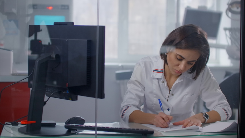 A female doctor sits at a table and writes with a pen writing out a prescription to a patient, behind a glass wall. Writing with a pen close-up Royalty-Free Stock Footage #1053766973