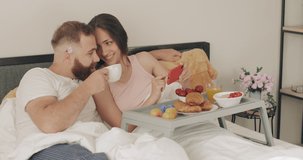 Happy young couple talking and smiling while looking at phone screen. Man drinking coffee while woman holding smartphone during breakfast in bed. Concept of relationship and leisure