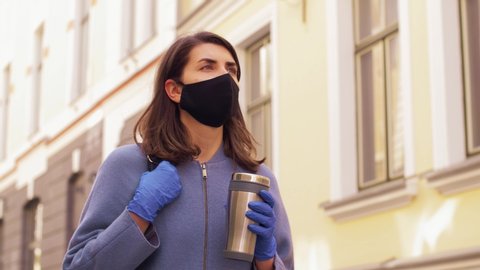 health, safety and pandemic concept - young woman wearing black face protective reusable barrier mask with tumbler or thermo cup walking in city