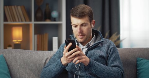 Young bearded guy in casual clothing sitting on couch and using smartphone with headphone for listening favorite music. Concept of relaxation and leisure time.