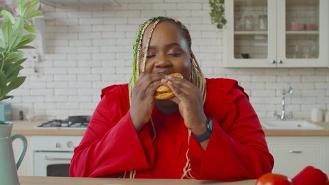 Charming hungry obese african american woman with afro braids enjoying eating harmful fast food, biting appetizing hamburger greedly, looking satisfied and happy in domestic kitchen.