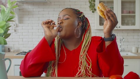 Beautiful hungry obese african female with afro braids on diet eating tasty hamburger in domestic kitchen, licking her finger with pleasure, avoiding healthy food and plan for weight loss.