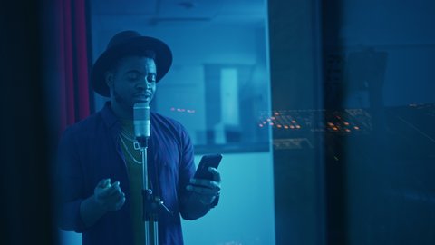 Portrait of Successful Young Black Artist, Singer, Performer Singing His Hit Song for the New Album. Wearing Stylish Hat, Holding Smartphone and Standing in Music Record Studio Soundproof Room,