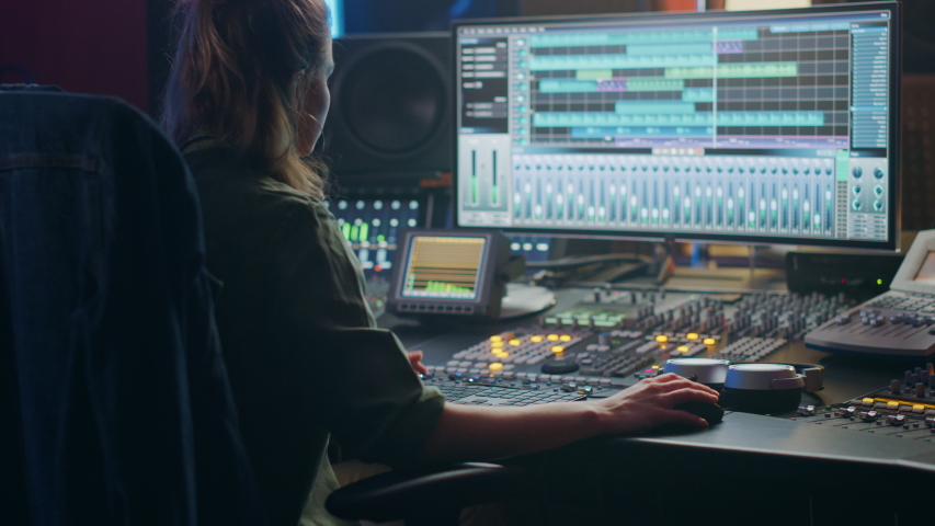 Beautiful, Stylish Female Audio Engineer and Producer Working in Music Recording Studio, Uses Mixing Board and Software to Create Cool Song. Creative Girl Artist Musician Working. Back View Zoom in Royalty-Free Stock Footage #1053781700