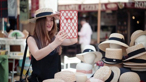 Amazing craft market full of different handmade products, pretty girl with adorable smile trying on stylish summer straw hat and looking into the mirror. Choice between hats styles on local flea