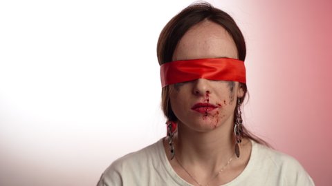 Tired woman with red ribbon on eyes sitting on white background with dropping blood from nose, scared female with beaten face is a symbol of freedom. Stop aggression against woman, violence concept