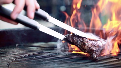 Roasting raw meat steak on burning fire, person using cooking tongs for turning roasted steak on bbq grid. Barbecue smoke and flames, grilling meat on fire. Barbecue and picnic concept