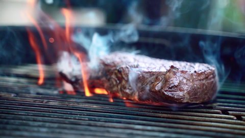 Close-up of fiery grilling steak on metal barbecue grid, roasting meat with spices and herbs for picnic party on terrace. Grilling juicy meat steak on burning charcoals with flames and smoke. Process