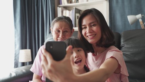 Asian family kid, mother, grandmother selfie photo and video by smartphone camera together in living room at home with happiness and smile. Happy activity technology lifestyle mobile phone use concept