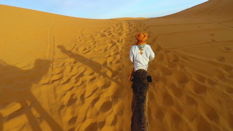 Man in traditional Berber robes leads a camel with tourist. Point of View of a ride of camel in sand dunes in the desert. Morocco