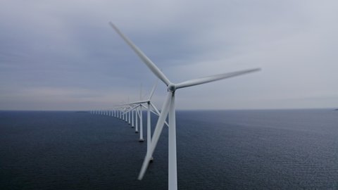 Panning shot from left to right of a row of windmills surrounded by deep sea waters, revealing an industrial area as the camera pans toward the left.