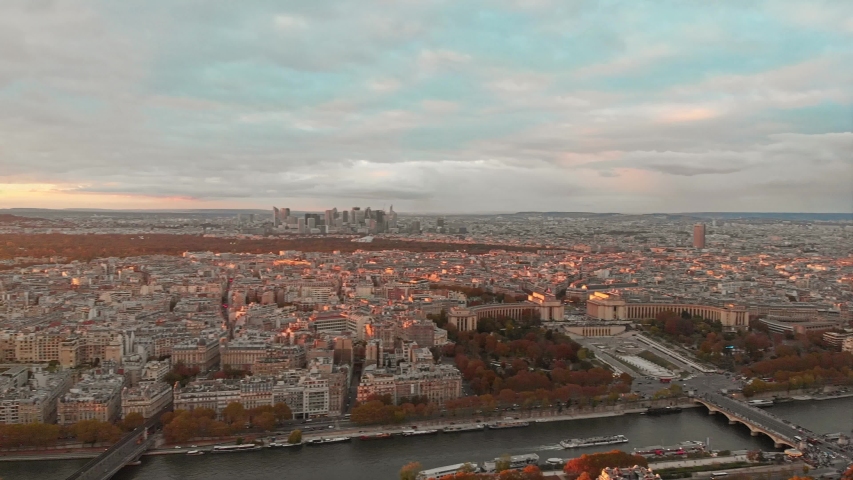 Aerial view of the Eiffel Tower and Trocadéro Gardens in Paris at sunset. The shot captures the Seine River, Palais de Chaillot, and the city skyline under a cloudy evening sky