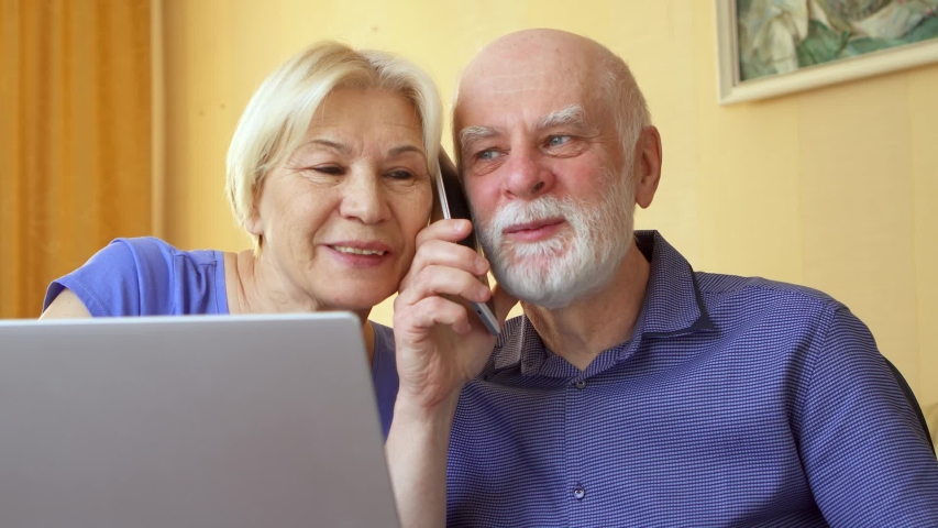Senior couple working from remote home office on laptop. Retired family staying home and using mobile phone to chat with their children. Active modern lifestyle on retirement | Shutterstock HD Video #1053795875