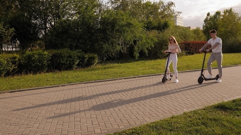 Fashionable young teenagers ride modern electric scooters in the Park in warm weather at sunset. Slow motion. A girl with long hair rides an electric scooter.