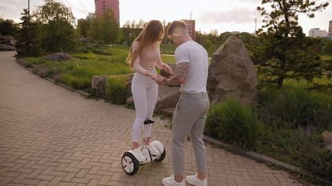 A young couple riding a hoverboard in a park in summer, self-balancing scooter. A guy teaches a girl to ride a gyro scooter. Slow motion.