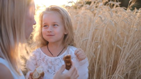 Mother with daughter eat a muffin for a walk in a wheat field. Mom and daughter have fun outdoors in the summer.