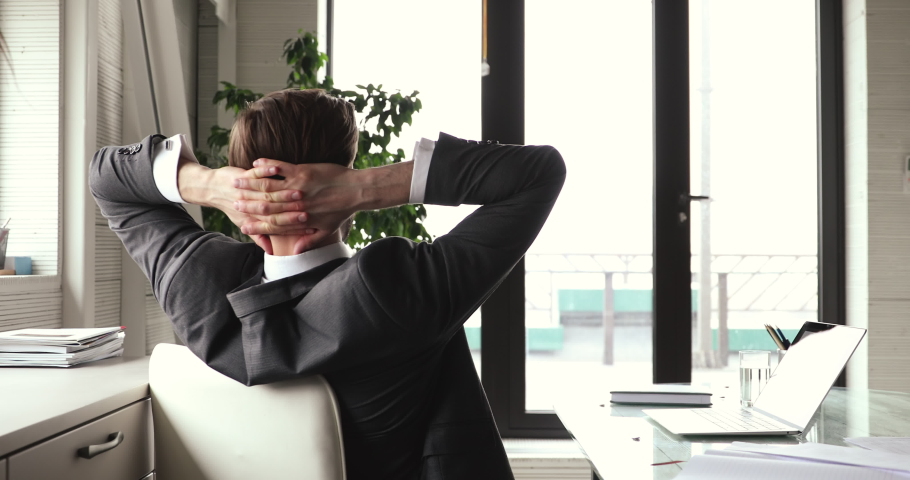 Happy young businessman wears suit taking break leaning on comfortable ergonomic office chair. Relaxed male executive puts hands behind head at workplace feels no stress after computer work well done. Royalty-Free Stock Footage #1053810857
