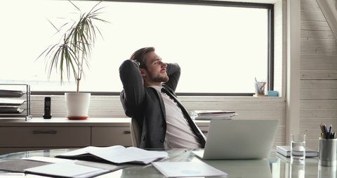 Successful satisfied young male company director wearing suit relaxing at workplace taking break after finishing computer work, chilling on chair, dreaming, feels peace of mind puts hands behind head.