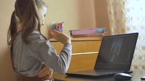 Back view of little girl with two ponytails saying hello for her tutor using video web camera on black laptop at home because of the self-isolation due to Coronavirus Covid-19. Remote education
