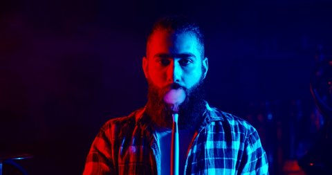 Authentic bearded guy smoking a hookah, exhaling clouds of white smoke in red and blue light - nightlife, niccotine addiction concept 4k footage close up portrait