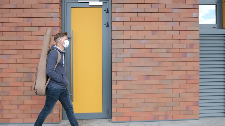 A young man with guitar in the back and with a medical mask on face is walking alone on the street. Social distancing. Pandemic situation