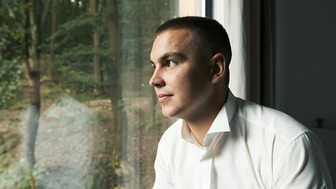 Young man of Caucasian appearance with brown eyes sitting by a panoramic window and thinking of a business plan